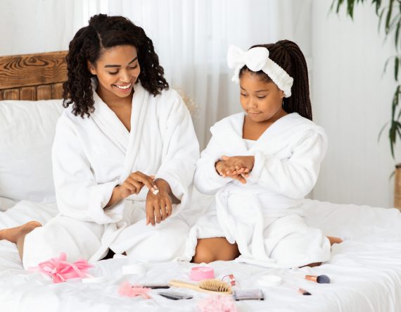 Mother Daughter Beauty Treatments. Caring Black Mom Teaching Little Girl Self-Care At Home, Sitting In Bathrobes On Bed, Applying Body Lotion On Hands, Different Makeup Cosmetics Lying Around